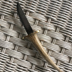 Puede incluir: Un pequeño cuchillo negro con patrón, con mango de asta de ciervo marrón y detalles dorados. El cuchillo está sobre una superficie de mimbre blanca.