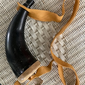 May include: A black powder horn with a wooden base and a leather strap. The horn is curved and has a small, round stopper. The leather strap is tan and appears to be made of suede.