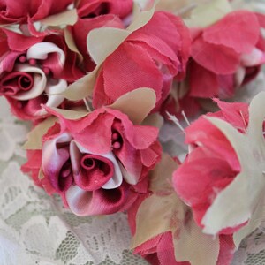 May include: A cluster of pink and white fabric roses with ruffled edges. The roses are arranged in a bouquet and are tied together with a white ribbon.