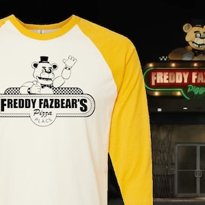 May include: A white and yellow baseball style long-sleeve shirt with a black and white graphic of Freddy Fazbear's Pizza. The graphic features a cartoon bear wearing a top hat and bow tie, waving. The text "Freddy Fazbear's Pizza Place" is written in a black banner.