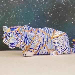 Può includere: Una statuetta di tigre bianca in posizione accovacciata, caratterizzata da strisce blu e linee color oro su tutto il corpo. La tigre presenta una superficie ruvida, orecchie a punta e una lunga coda che si estende dietro, in forma dettagliata.