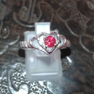 May include: A silver ring with a heart-shaped design and a round, red gemstone at the center. The ring is displayed on a clear stand, showcasing its intricate details and the vibrant color of the stone. The band has a textured pattern.