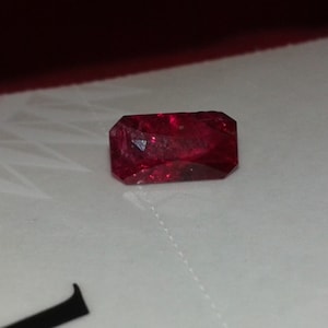 May include: A faceted, rectangular-cut, deep red gemstone. The gem has a brilliant, reflective surface and is set against a white background. The gem's shape is an elongated rectangle with cut corners.