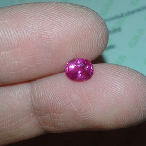 May include: A small, round, faceted, hot pink gemstone. The gem is held between two fingers, showcasing its brilliant cut and vibrant color. The gem is likely a ruby or pink sapphire.