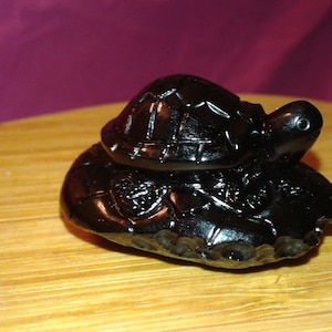 Puede incluir: Pequeña figura de tortuga oscura. La tortuga está tallada con patrones detallados en su caparazón y cuerpo. La figura está sobre una superficie de madera, con un fondo morado borroso.