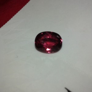 May include: A faceted, oval-shaped, deep red gemstone. The gem has a polished surface that reflects light, creating bright highlights. The stone is set against a white surface with a red fabric in the background.