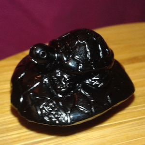 Puede incluir: Una figura de tortuga negra brillante sobre una base texturizada. La tortuga tiene un caparazón detallado y una cabeza pequeña con ojos visibles. La figura se encuentra sobre un fondo borroso.