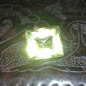 May include: A square-cut, light yellow gemstone with a central facet, resting on a dark, textured surface. The gemstone reflects light, creating a bright, glowing effect. The surface has an embossed pattern.