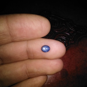 May include: A small, oval-shaped, blue gemstone. The gem is held between two fingers, with a dark background. The stone has a smooth, polished surface and reflects light, creating a subtle shine. The gem's color is a deep, saturated blue.