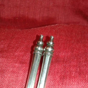 May include: Two silver-colored, elongated objects with decorative tops, possibly serving as serving utensils or decorative elements. The objects are set against a red fabric background, creating a contrast in color and texture.