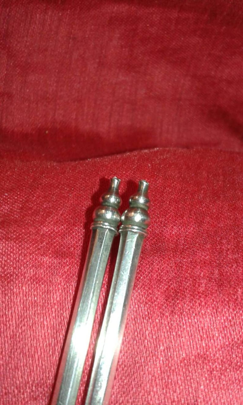 May include: Two silver-colored, elongated objects with decorative tops, possibly serving as serving utensils or decorative elements. The objects are set against a red fabric background, creating a contrast in color and texture.