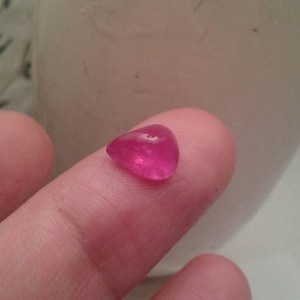 Op de afbeelding: Een gepolijste, druppelvormige, levendige roze edelsteen. De edelsteen wordt tussen twee vingers gehouden, waardoor het gladde, reflecterende oppervlak wordt getoond. De achtergrond is een vage, gebroken witte ondergrond.