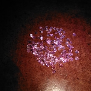 May include: A collection of small, round, faceted gemstones in varying shades of pink and purple. The gems are scattered on a dark brown surface, creating a contrast of color and texture. The gemstones appear to be of high quality.