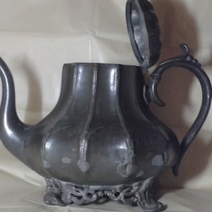 Puede incluir: Una tetera de peltre con un pico y asa curvados, y una tapa con bisagras. La tetera tiene una base decorativa y un cuerpo acanalado. La tapa está abierta, revelando el interior. La tetera es de color gris oscuro.