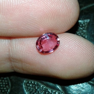 May include: A faceted, oval-shaped gemstone with a pinkish-red hue. The gem is held between two fingers, showcasing its polished surface and internal clarity. The gem's color is consistent throughout, with a slight variation in tone.