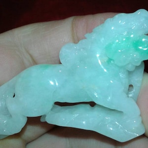 May include: A carved jade figurine of a mythical creature, possibly a lion or dragon. The sculpture is primarily a pale green color with areas of brighter green. The figure is held in a hand.