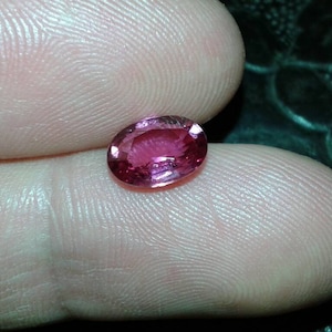May include: A vibrant, oval-cut, pink gemstone. The gem is held between two fingers, showcasing its polished surface and internal facets. The stone's color ranges from deep rose to a lighter pink, reflecting light.