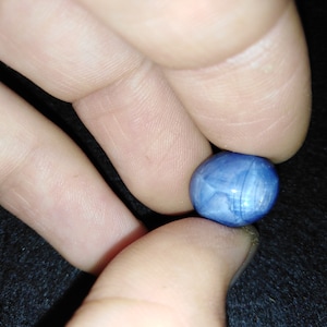 May include: A small, round, blue gemstone with a polished surface. The stone is held between two fingers, showcasing its color and texture. The gemstone appears to be a cabochon, a style of cutting that emphasizes the stone's color and pattern.