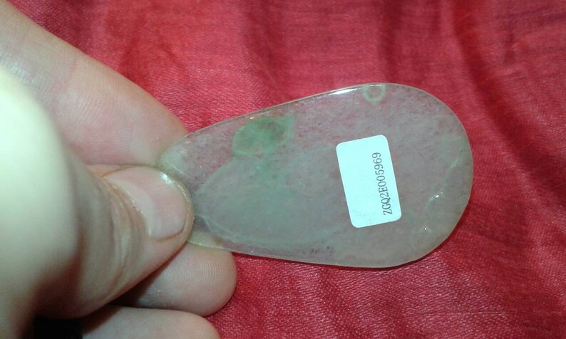 May include: A translucent, teardrop-shaped pendant with a pale green hue and a small, attached label. The pendant has a subtle, cloudy appearance and a small hole at the top. The label displays the text "ZCQ2E005969".