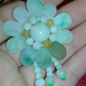 May include: A floral pendant necklace featuring a flower design made of light green and white jade stones. The pendant has a central large round stone, surrounded by petal-shaped stones, with smaller beads and teardrop-shaped stones. The necklace has a black cord.