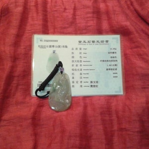 May include: A jade pendant with a black cord and a certificate of authenticity. The pendant is a light green color with a carved design. The certificate has Chinese characters.