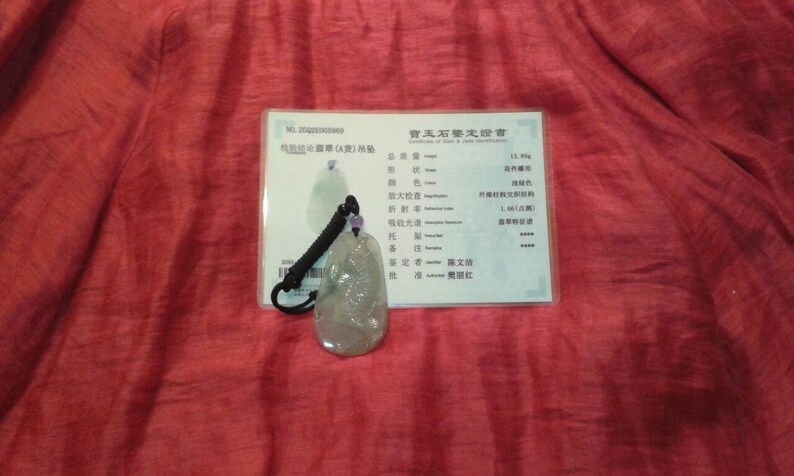 May include: A jade pendant with a black cord and a certificate of authenticity. The pendant is a light green color with a carved design. The certificate has Chinese characters.