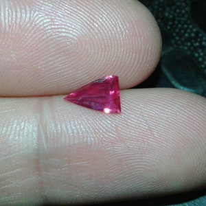 May include: A triangular, deep pink gemstone with multiple facets. The gem is held between two fingers, highlighting its vibrant colour and cut. The facets of the gem reflect light, creating a sparkling effect. This gemstone could be used in jewellery.