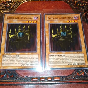 Pode incluir: Duas cartas colecionáveis com o texto "CASTELO DAS ILUSÕES SOMBRIAS" e uma ilustração de um castelo. As cartas têm um esquema de cores dourado e preto. Cada carta tem um ATK de 920 e DEF de 1930.