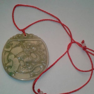 May include: A round, pale green jade pendant with a carved design, suspended on a bright red cord necklace. The pendant features a detailed relief carving. The red cord is knotted and has small beads.