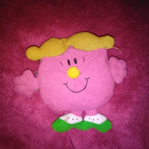Puede incluir: Un peluche rosa con pelo amarillo, una nariz amarilla y una cara sonriente. El juguete tiene brazos pequeños y zapatos verdes y blancos. El juguete está sobre un fondo rosa.