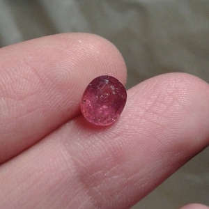 Puede incluir: Una pequeña gema translúcida de forma ovalada, con un tono rosa intenso. La gema se sostiene entre dos dedos, mostrando su superficie pulida e inclusiones naturales. El color de la gema varía de rosa claro a oscuro.