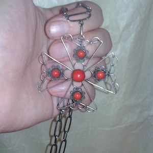Könnte beinhalten: Silberfarbene Kreuz-Anhänger-Halskette mit Kette. Das Kreuz hat ein filigranes Design mit fünf roten, korallenartigen Steinen. Die Halskette wird in einer Hand gehalten.