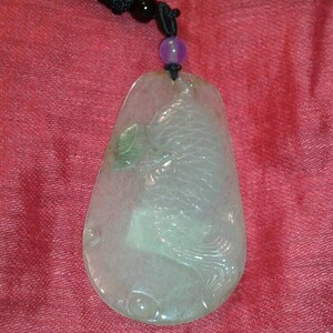 May include: A translucent jade pendant with a carved fish design. The pendant is teardrop-shaped and hangs from a black cord with a purple bead. The jade has a subtle green hue and is set against a red background.
