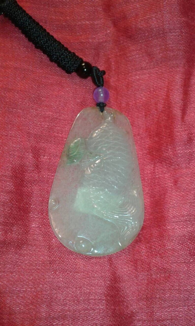 May include: A translucent jade pendant with a carved fish design. The pendant is teardrop-shaped and hangs from a black cord with a purple bead. The jade has a subtle green hue and is set against a red background.