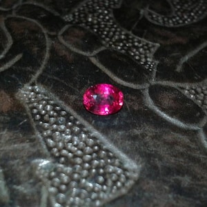 May include: A vibrant, oval-cut, hot pink gemstone rests on a dark, textured surface. The gemstone's facets catch the light, displaying a brilliant sparkle. The background features an intricate, embossed pattern.
