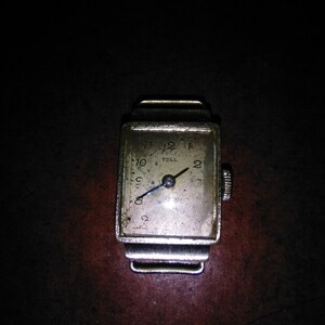 Puede incluir: Un reloj de pulsera rectangular vintage con caja plateada y esfera desgastada blanquecina. El reloj tiene números negros, manecillas negras y la palabra "TELL". El fondo es oscuro.