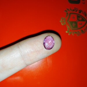 May include: A faceted, oval-shaped, purple gemstone. The gem is held on a finger against a bright orange background. The gem's facets catch the light, creating a sparkling effect. The image is well-lit, highlighting the gem's color and clarity.