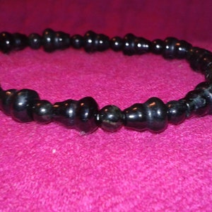 May include: A black beaded bracelet with a glossy finish. The beads have a unique, rounded shape, and the bracelet is displayed on a pink surface. This jewelry item is suitable for everyday wear.