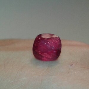 May include: A faceted, cushion-cut ruby gemstone. The gem is a deep, saturated red color with internal inclusions, and is displayed against a neutral background.