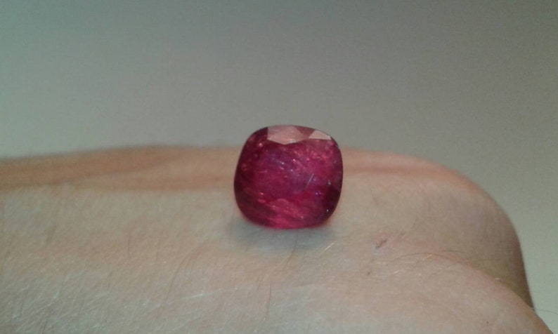 May include: A faceted, cushion-cut ruby gemstone. The gem is a deep, saturated red color with internal inclusions, and is displayed against a neutral background.