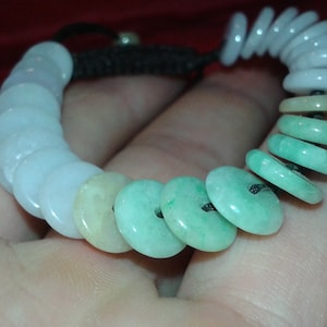 May include: A jade bracelet with disc-shaped beads in white, green, and yellow hues. The beads are strung on a black cord. The bracelet is a stylish accessory, with the beads likely measuring around 1-2 cm in diameter.