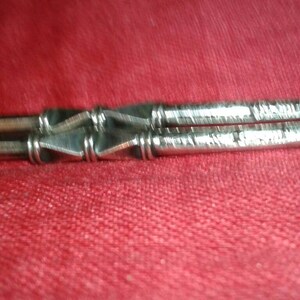May include: Close-up of a silver metal object with a geometric design. The object appears to be two parallel rods connected by a series of diamond-shaped elements. The rods have etched lines. The background is a solid red color.