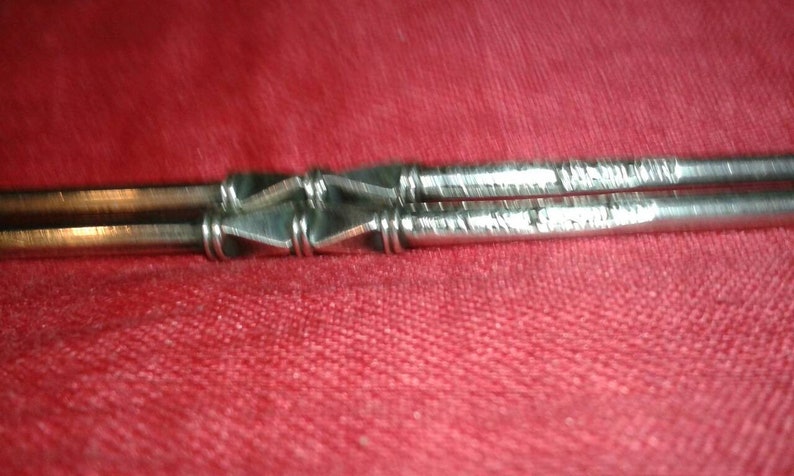 May include: Close-up of a silver metal object with a geometric design. The object appears to be two parallel rods connected by a series of diamond-shaped elements. The rods have etched lines. The background is a solid red color.