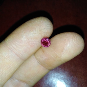 May include: A small, round, faceted gemstone with a vibrant pink hue. The gem is held between two fingers, showcasing its size and color against the skin. The gem's surface reflects light, highlighting its clarity and brilliance.