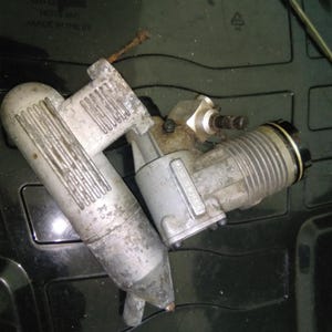 May include: A used, silver-colored model engine with a cylindrical body and a curved exhaust pipe. The engine has visible wear and tear, with some rust and discoloration. The text "MADE IN RUSSIA" is visible on the engine.