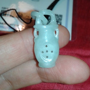 May include: A pale green jade pendant carved as a small vase with a looped handle and decorative holes. It hangs from a black cord. The background features a red surface and a white card. The pendant is likely a decorative item.