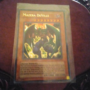 May include: A Yu-Gi-Oh! trading card featuring "Mazera DeVille" in a protective sleeve. The card has a gold background with a monster illustration. Text includes "LIMITED EDITION" and card details.
