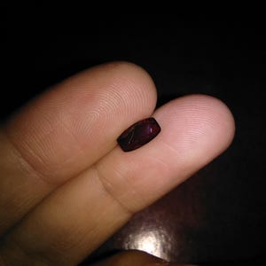 May include: A small, rectangular, dark red gemstone is held between two fingers. The gem has a polished surface and appears to be faceted. The background is a dark, solid colour, emphasising the gem's colour and shape.