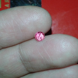 Può includere: Una piccola gemma rosa e rotonda, forse un rubino o uno zaffiro rosa, è tenuta tra due dita. La gemma è di un colore rosa vibrante e saturo e sembra sfaccettata. Lo sfondo è rosso pieno.