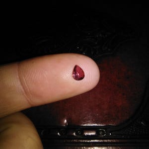 Puede incluir: Una pequeña gema roja oscura en forma de lágrima descansa sobre la yema de un dedo. La gema está muy pulida y refleja la luz, sobre un fondo oscuro. La imagen resalta el color y la forma de la gema.